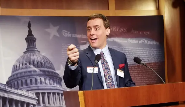 A man in a suit and tie delivers a speech at a podium, engaging the audience with his presentation.