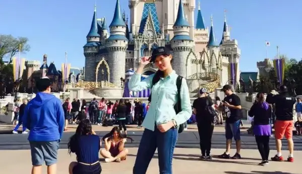 A woman wearing a blue shirt poses in front of a grand castle, showcasing its impressive architecture.
